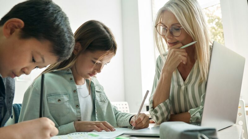 Female teacher helping diverse kids students with homework in classroom.
